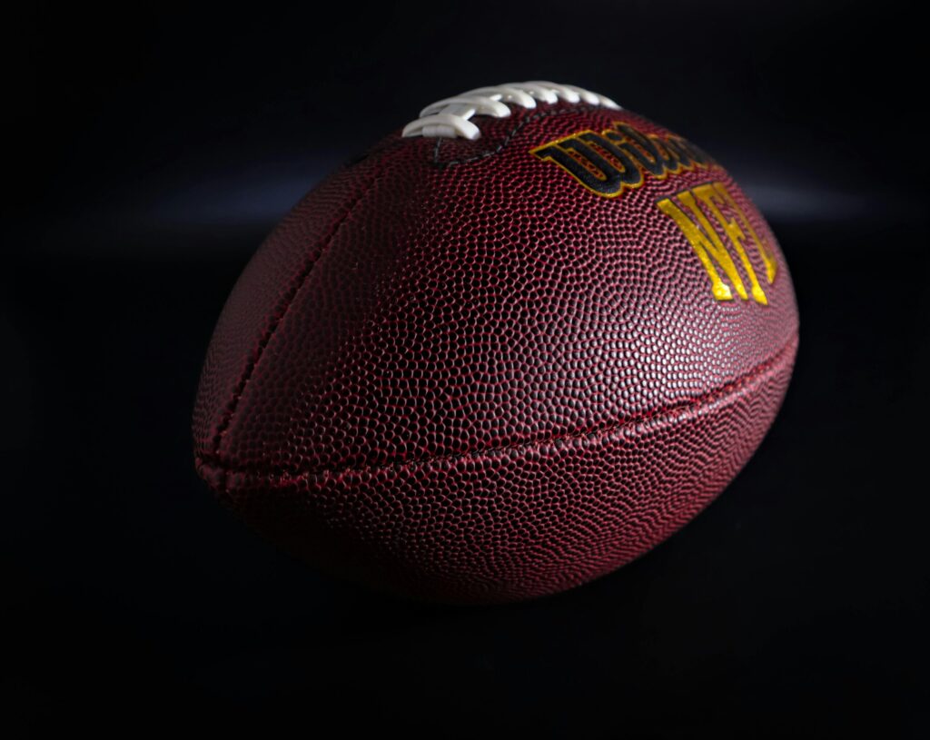 3 Games that Will Shape the NFL Playoff Picture Detailed view of a traditional brown and white American football against a dark backdrop, highlighting texture.