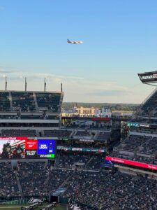 A plane soars above a bustling football stadium, capturing the excitement of live sports under a blue sky.