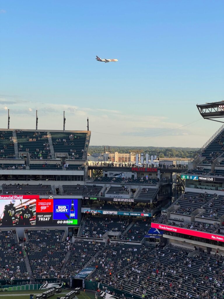 A plane soars above a bustling football stadium, capturing the excitement of live sports under a blue sky.